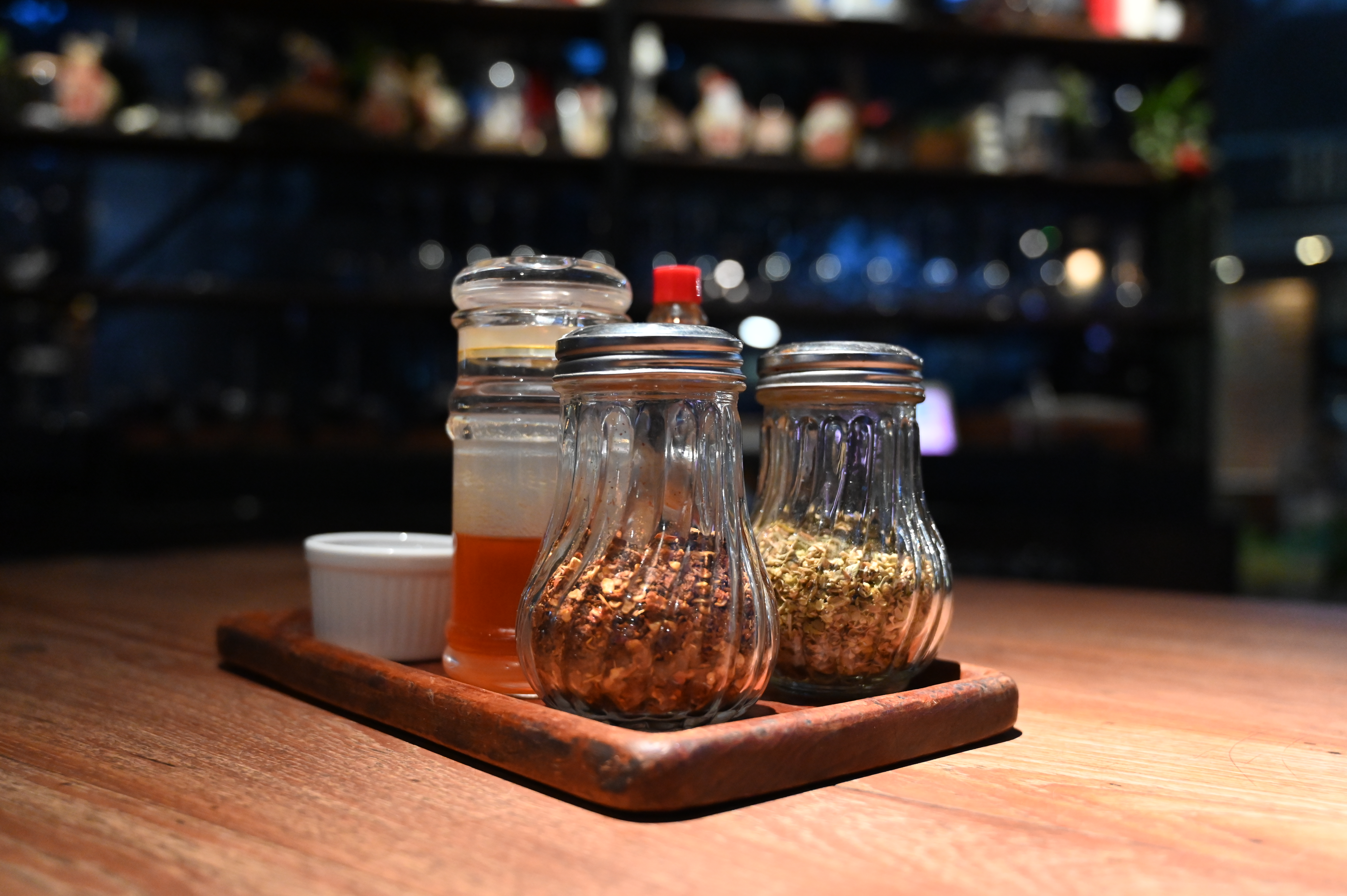 Glass shakers of chili flakes and herbs with a jar of sauce on a wooden tray