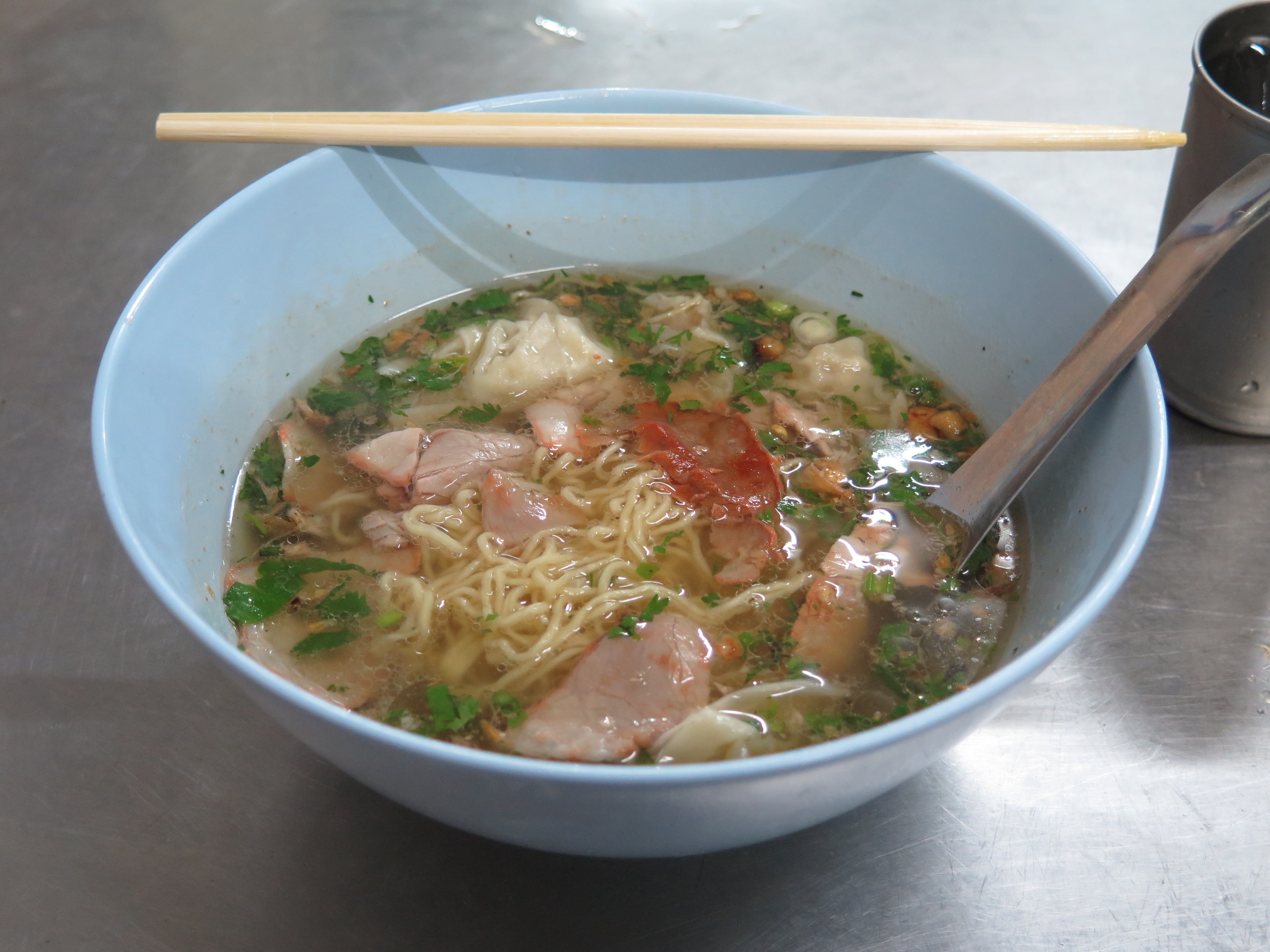 Bowl of noodle soup with dumplings and sliced pork on a steel counter