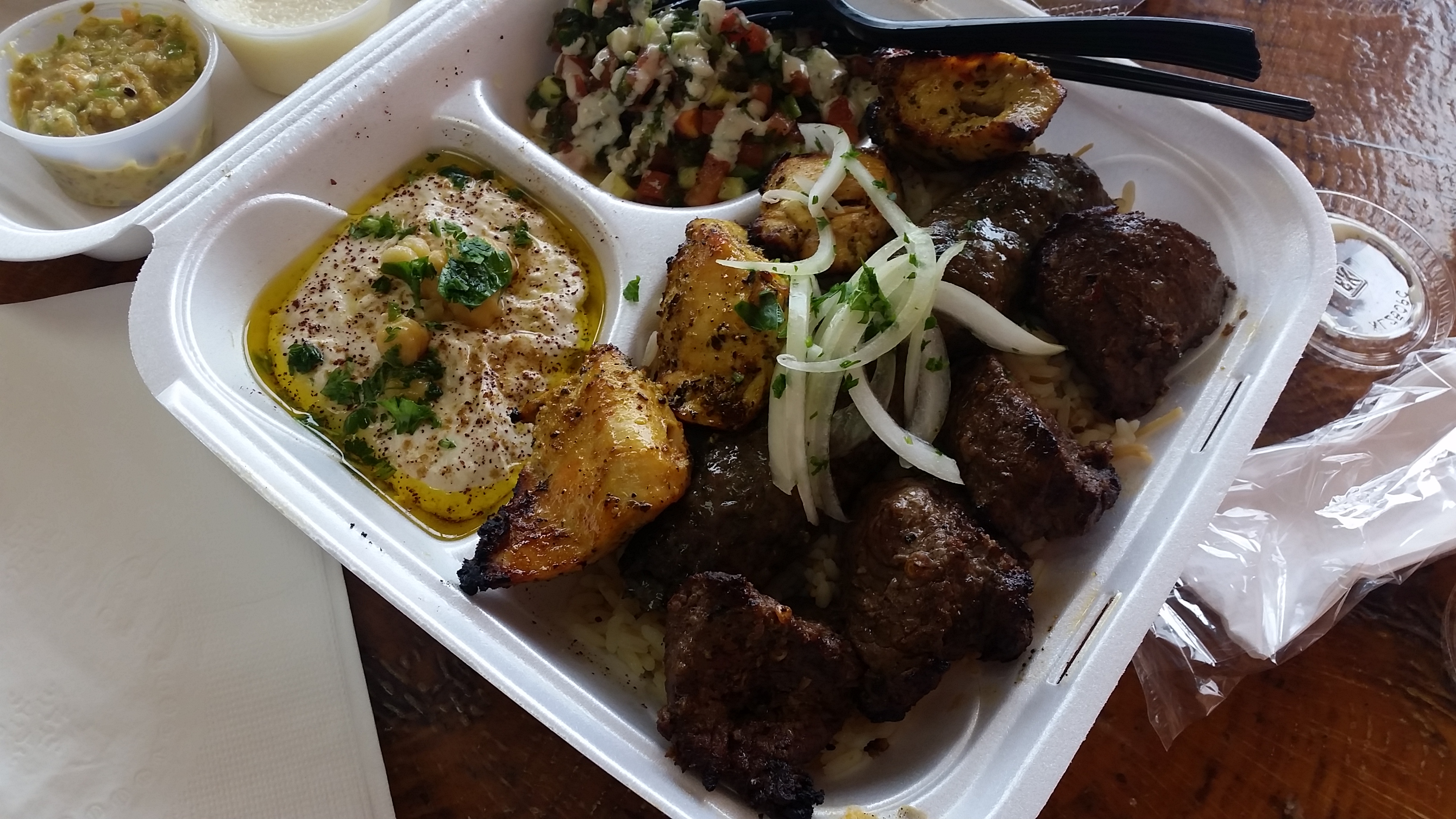 A delivered takeout tray of grilled kebabs, hummus, rice and salad with side pots and plastic cutlery