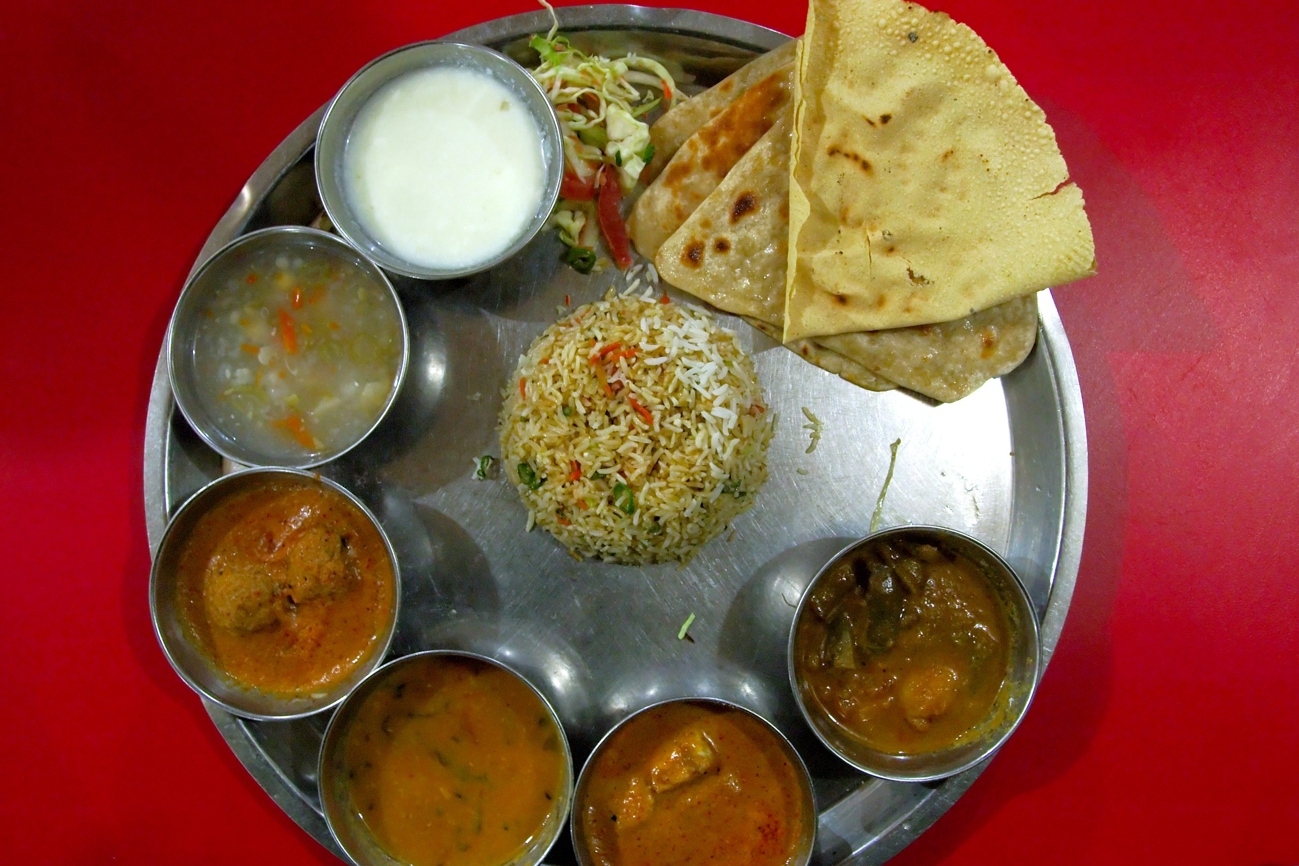 Steel thali plate with rice, flatbreads, curries in small bowls and a cup of yogurt