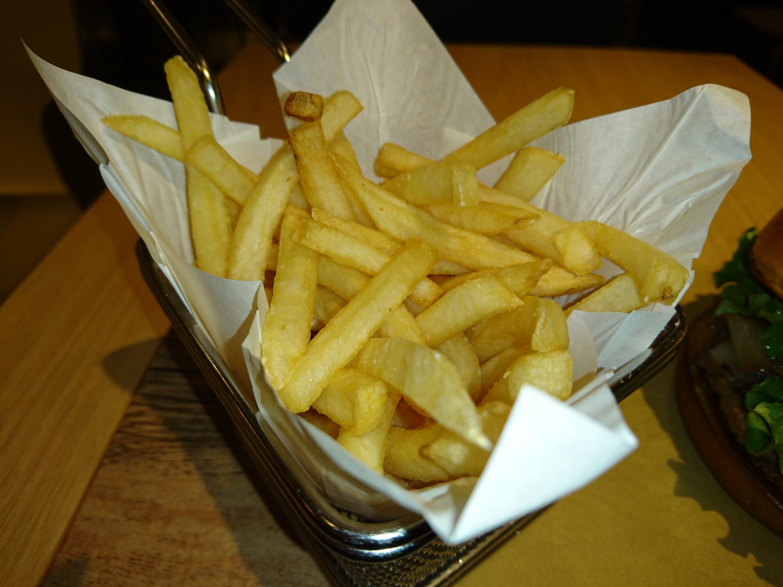 Wire basket of french fries lined with paper next to a burger on a wooden table