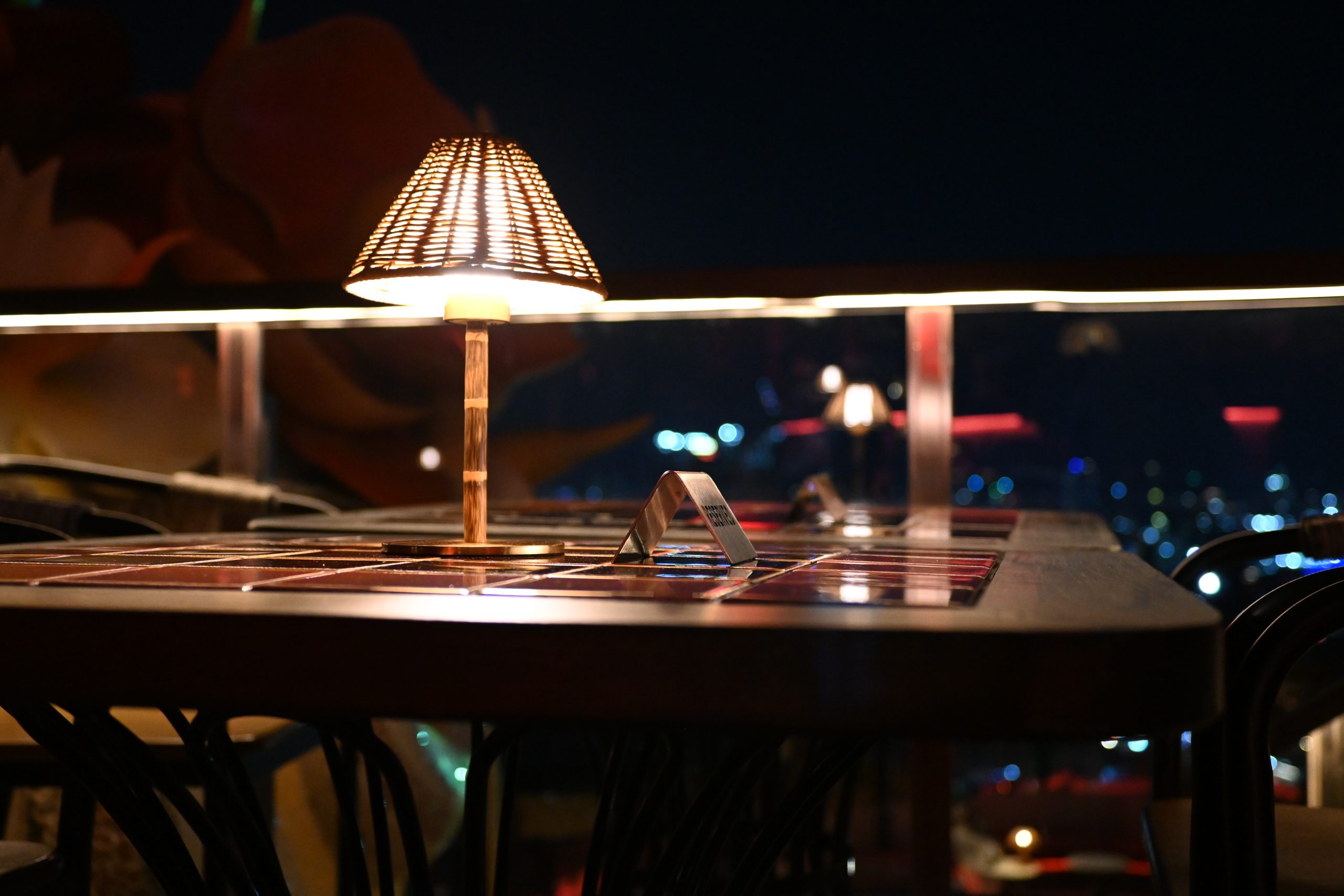 Restaurant table lit by a small woven lamp at night with city lights in the background