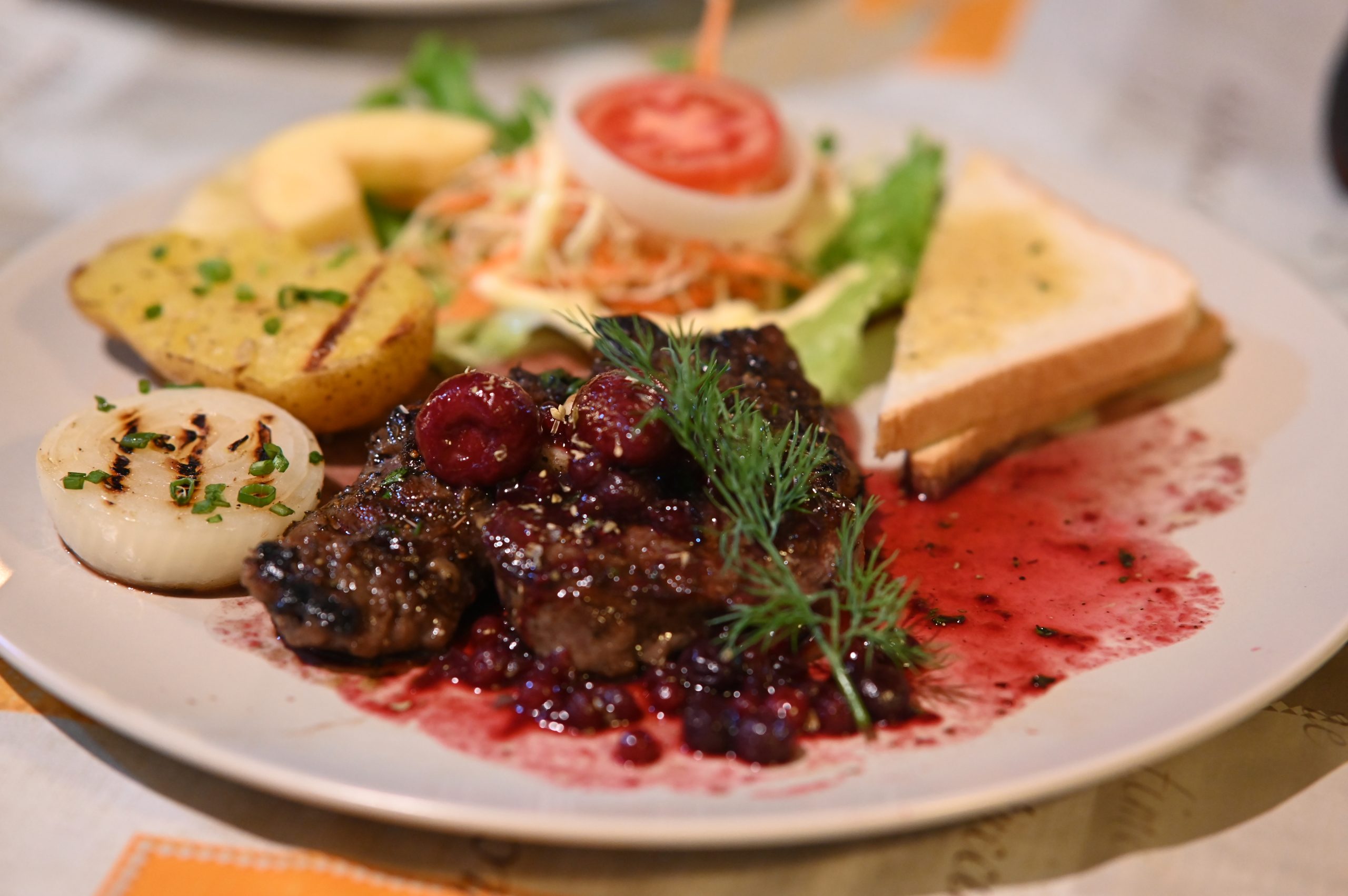 Seared steak medallions with berry sauce, roasted potato, grilled onion, toast and side salad on a white plate