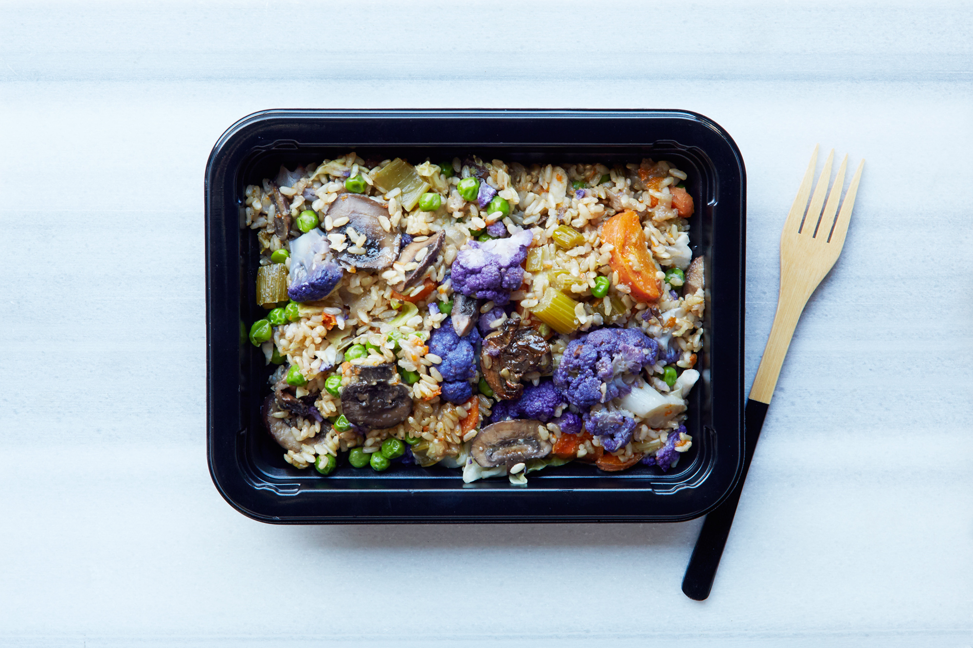 Black meal prep container of brown rice with peas, mushrooms, carrots and purple cauliflower beside a wooden fork