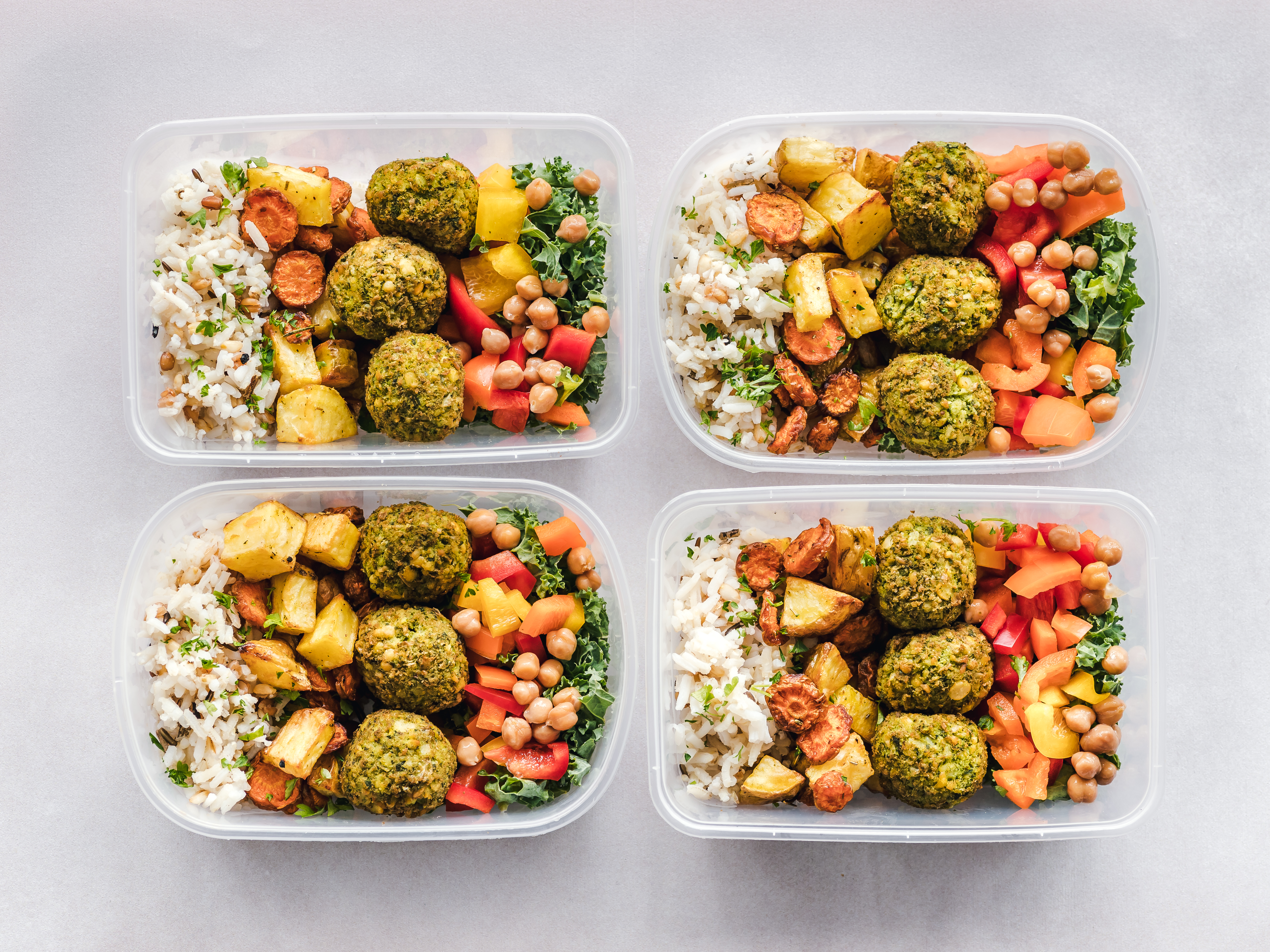 Four plastic meal prep boxes with rice, falafel, roasted vegetables and chickpeas