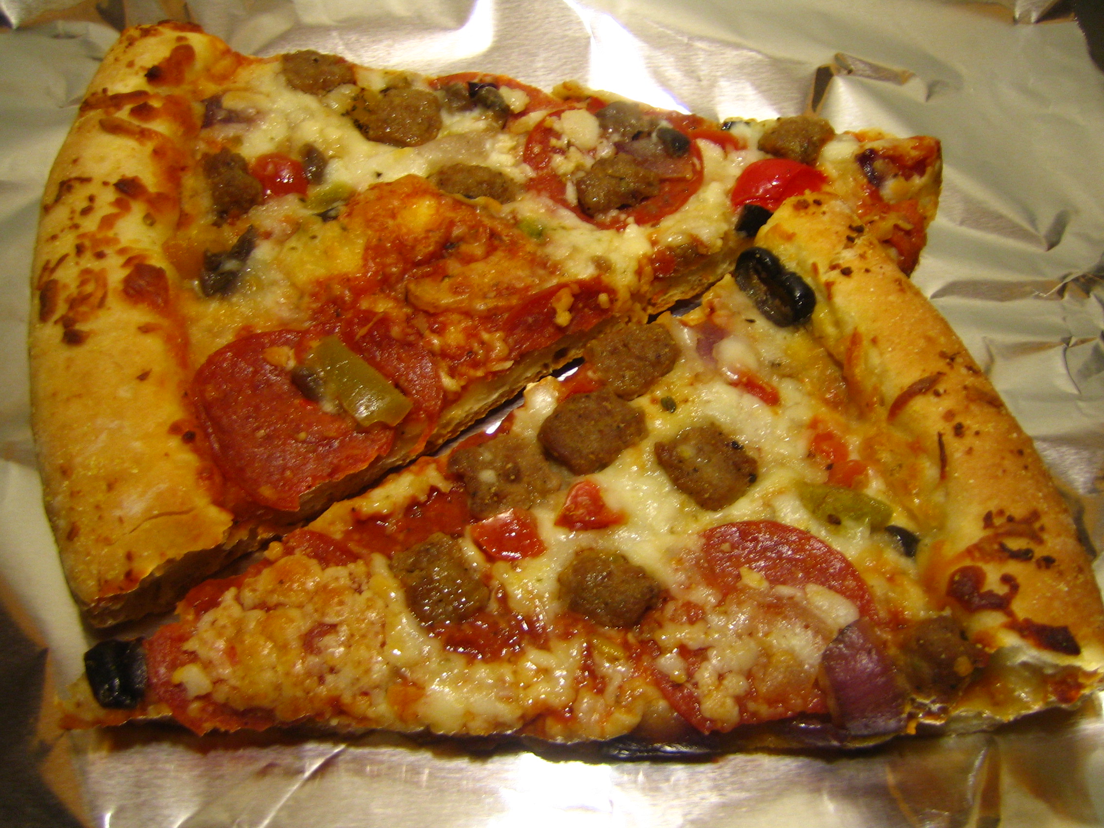Two slices of leftover pizza with sausage, pepperoni and olives on aluminum foil