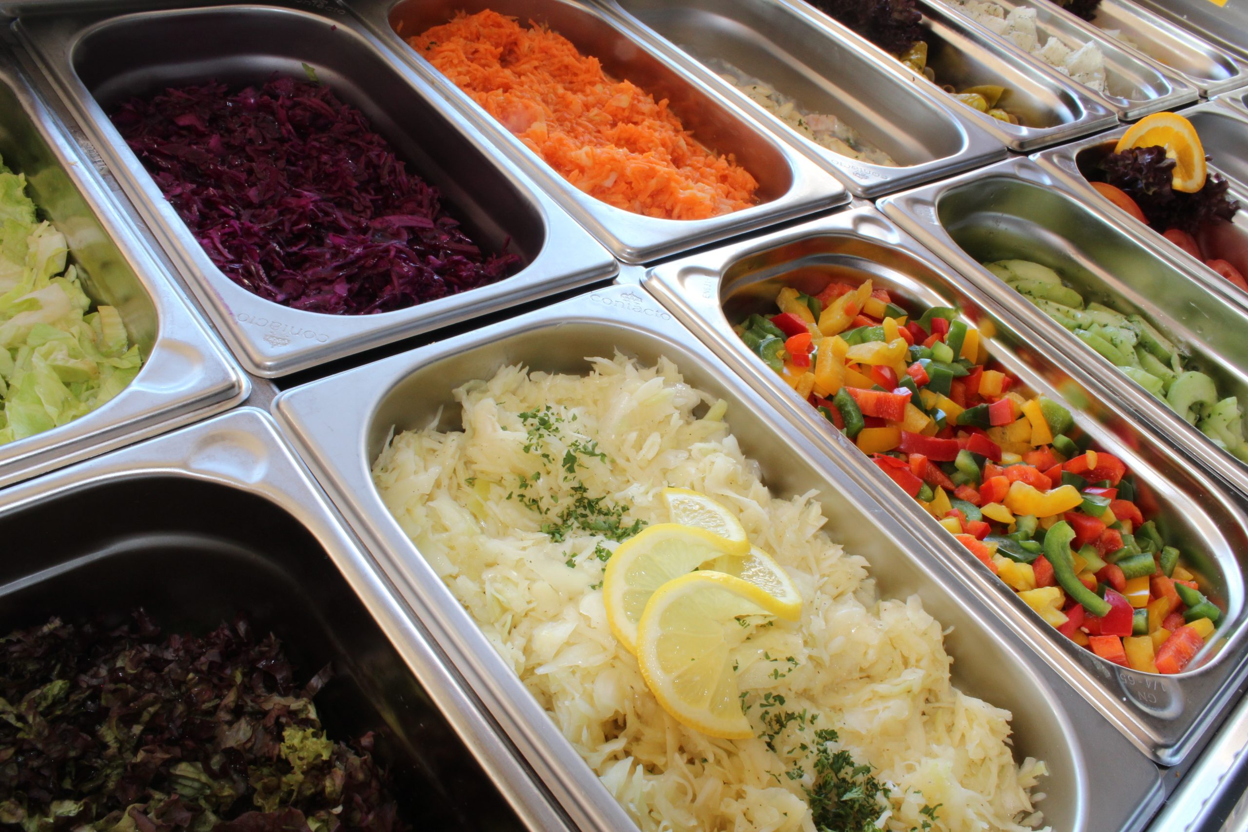 Steel trays of shredded cabbage, red cabbage, grated carrot, peppers and cucumber at a salad bar